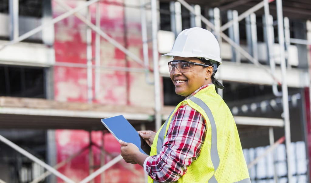 A mature African-American woman in her 40s working at a construction site, wearing a hardhat, safety goggles and reflective vest. She is looking over her shoulder at the camera with a confident expression, smiling, holding a digital tablet. A building under construction is out of focus in the background.