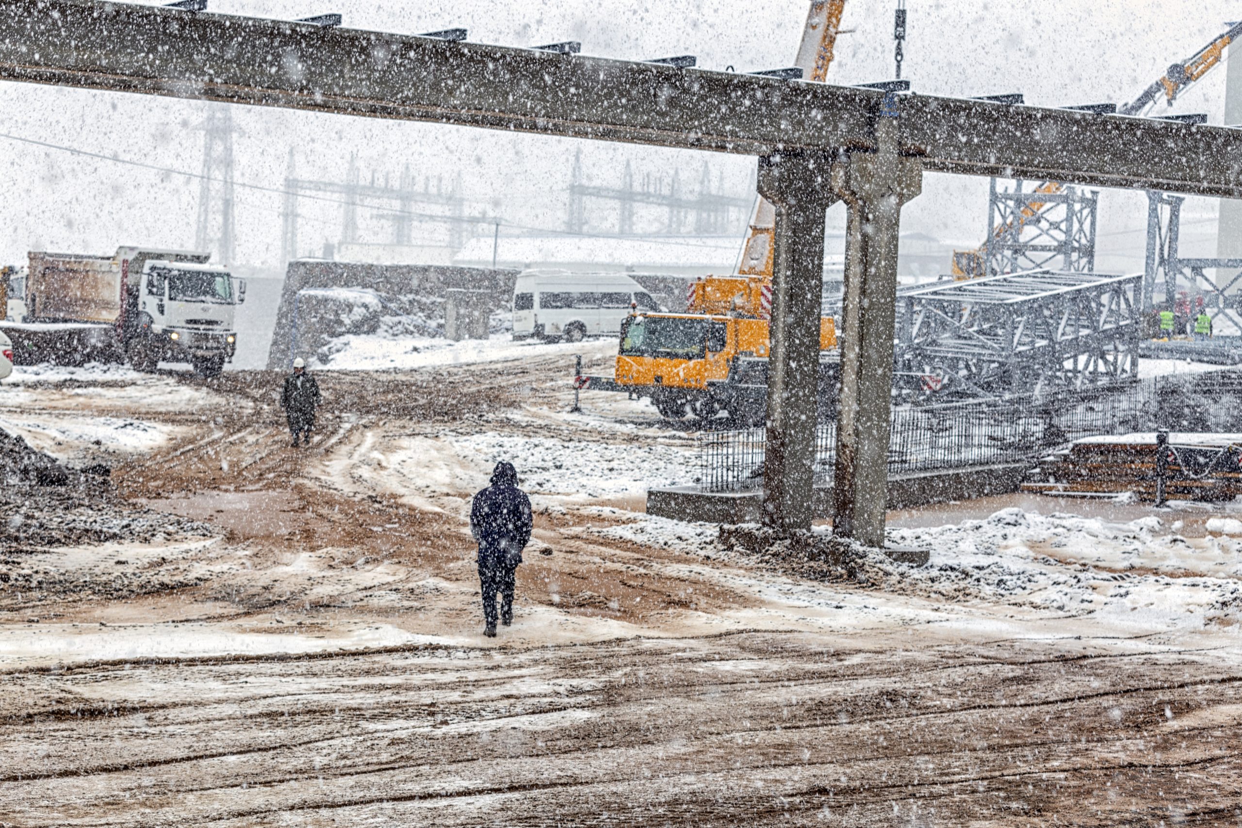 A man at a winter construction site with a rail brdge abovee.