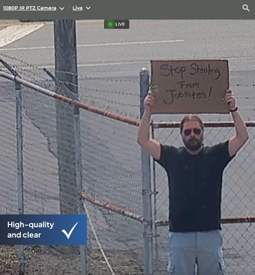 An image of a man standing in a parking lot holding up a sign that says "Stop Stealing form Jobsites!" captured by a construction camera that is zoomed in to read the sign.
