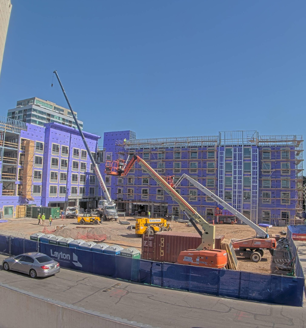 Hilton Park Central hotel under construction with three aerial lifts on the construction site and a few workers wearing PPE. The building has purple sheathing on it and a fence around it 