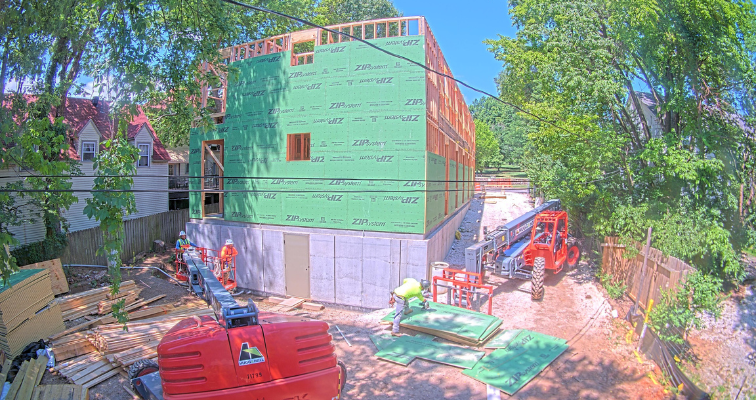 Construction site with a building in the framing stage with ZIP System sheathing panels installed and construction workers installing more. Captured by a construction camera