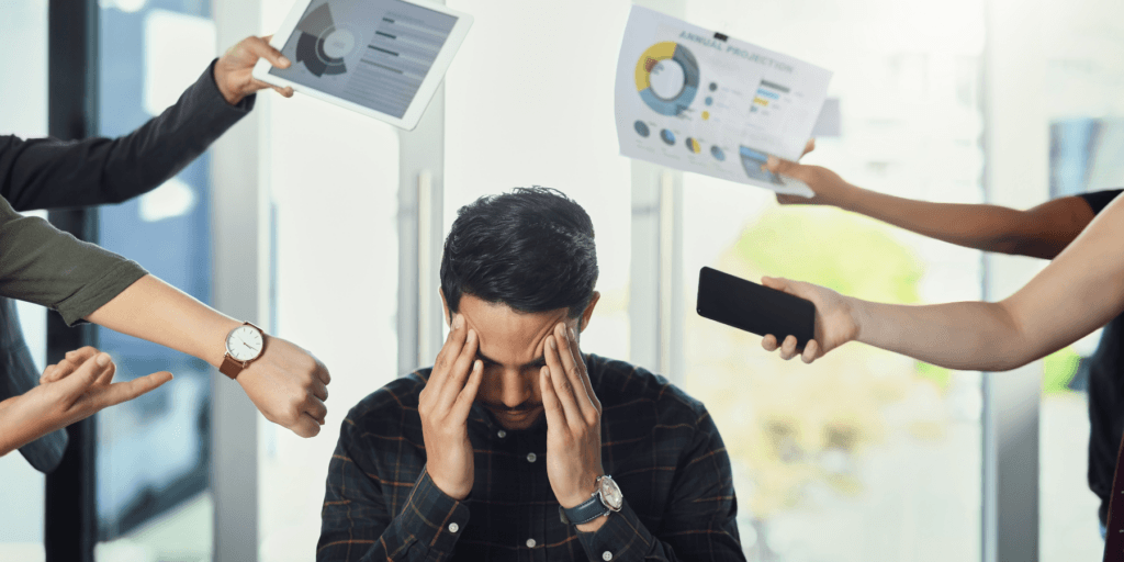 A stressed man with hands on his temples while there are five other hands above him handing him presentations, phones, and pointing at a watch