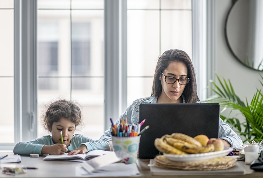 Woman working on a laptop at home while a child draws beside her