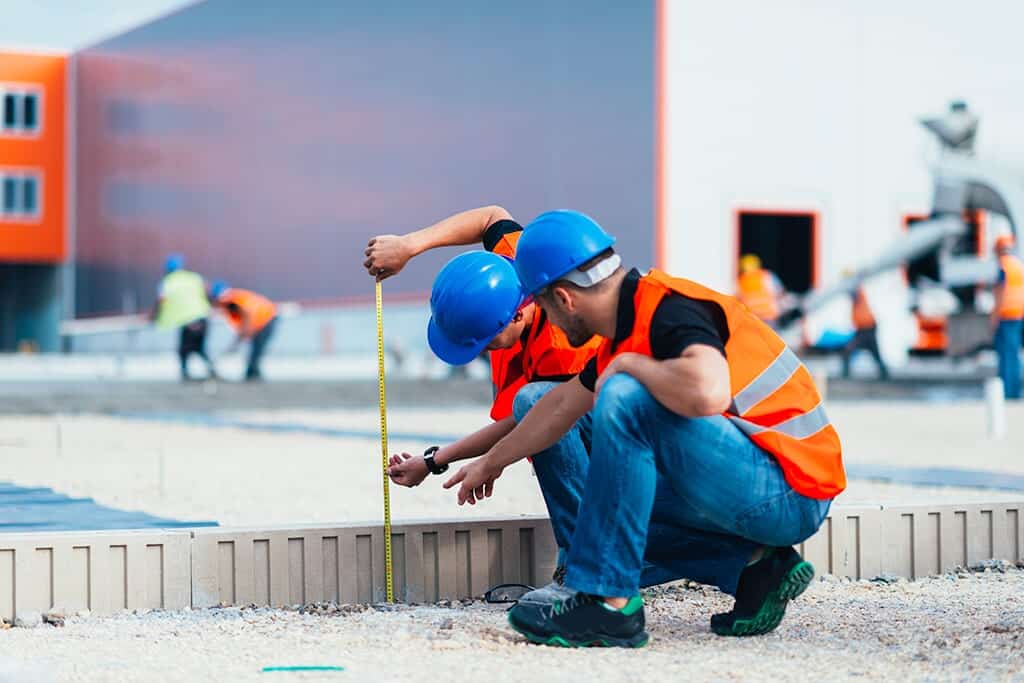 Two construction workers in hard hats measuring a concrete form with a tape measure