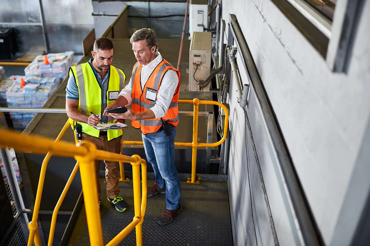 Two workers in safety vests reviewing a tablet in a warehouse