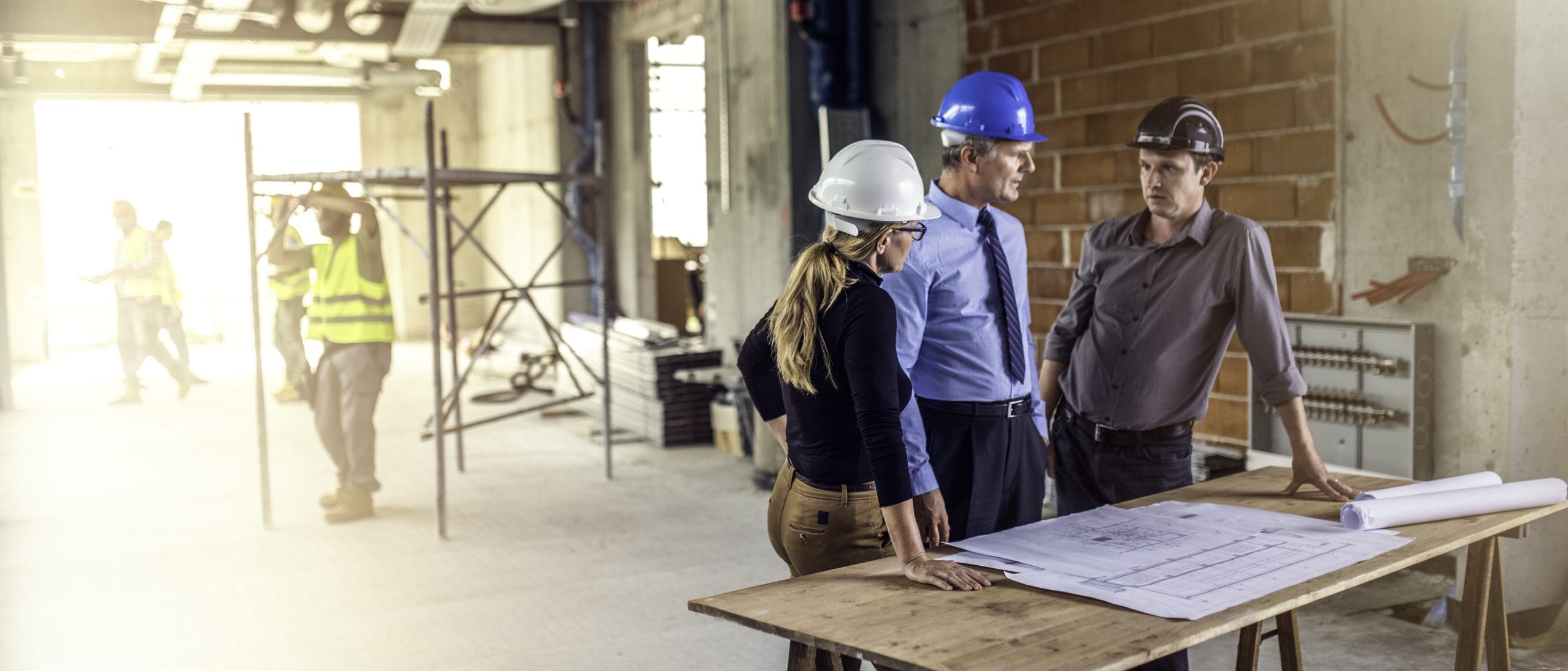 Two male project managers in hard hats with a female project manager standing around a table with a blueprint on it at a construction site while subcontractors do work in the background