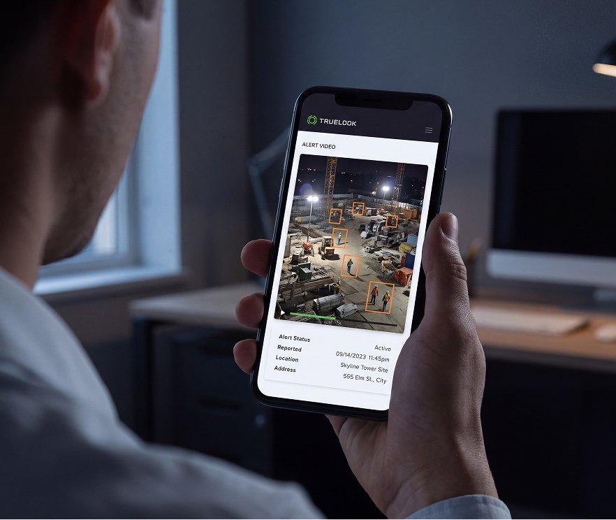 Person holding a smartphone showing a TrueLook alert video of a jobsite at night